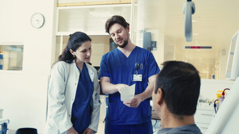 Eine Ärztin im weißen Kittel und ein Pfleger in blauer Kleidung besprechen medizinische Unterlagen mit einem Patienten.